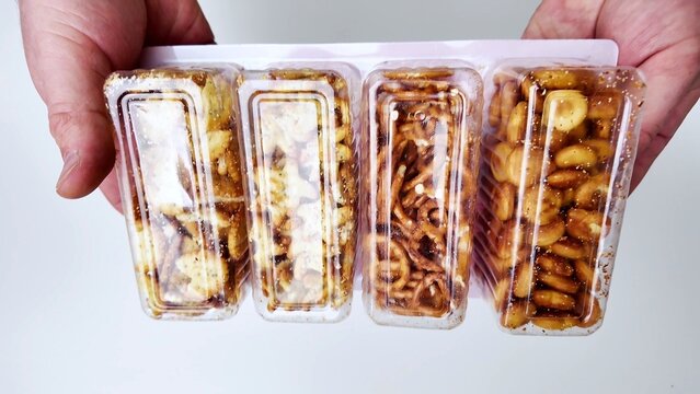 A person holds a plastic package containing four separate compartments of assorted salty snacks, including pretzels and round crackers, against a white background