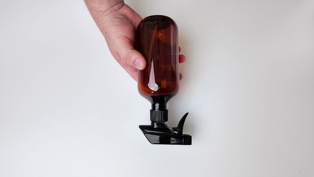 A person's hand gently holds a dark amber colored glass bottle containing a thin stalk, positioned on a plain white surface, presenting a focus on the object and skin tones