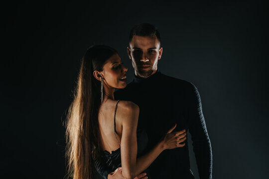 A stylish couple embraces in a dark studio setting, the woman wearing a backless dress and long earrings. Moody lighting highlights their intimate pose and fashionable evening wear.