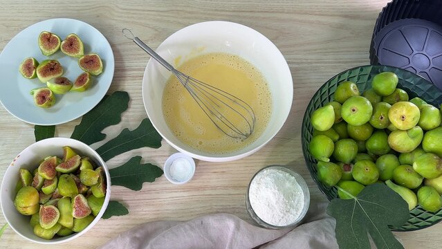 green figs in a bowl and sliced figs on a plate surround a bowl of yellow batter with a whisk, flour in a glass cup, and a small dish of powder on a wooden table