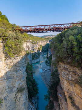Aerial view of Osumi canyon featuring a metal bridge spanning the deep rocky gorge with a turquoise river flowing below surrounded by lush greenery in Dhores, Berat County, Albania.