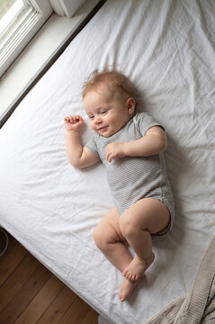 Cute infant baby lying on back on a white bed near window, top view. Happy adorable newborn in striped bodysuit smiling at home.