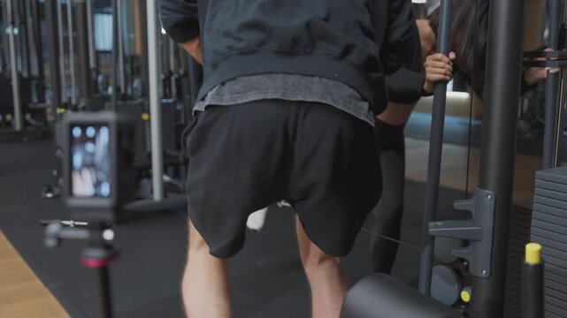 Male personal trainer coaches a pregnant client through cable glute kickbacks while an action camera records the session in slow motion.