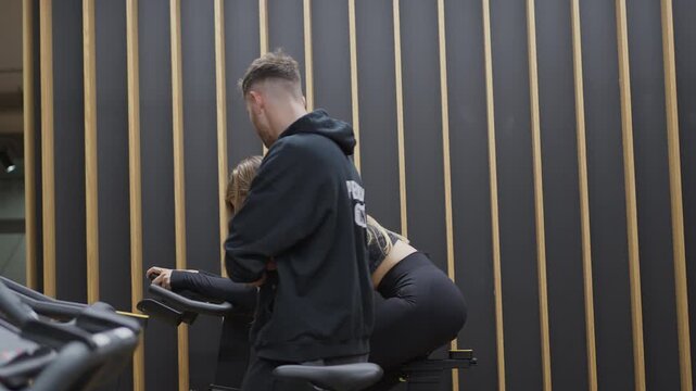 Smiling male personal trainer stands behind a woman pedaling a stationary bike, then walks toward her during a cardio session in slow motion.