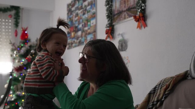 A grandmother lifts and hugs her laughing toddler grandchild in a living room decorated with a Christmas tree and tinsel garlands