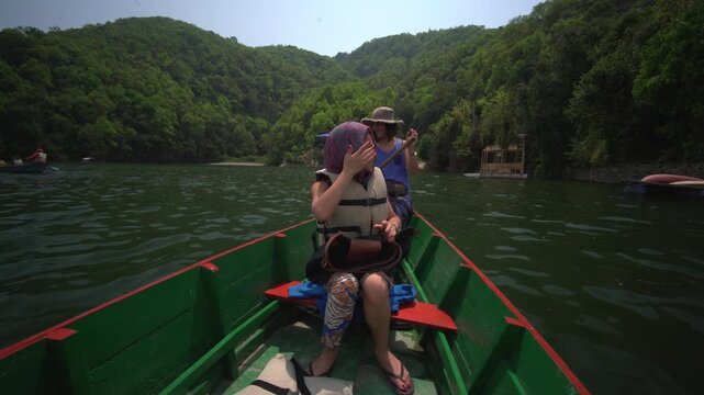 Two women in sun hats row a wooden boat along a narrow forested channel of Phewa Lake in Pokhara