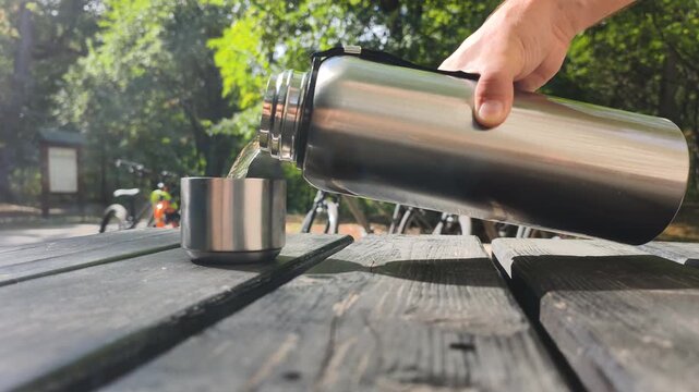 Outdoor picnic relaxation: pouring hot beverage from steel flask into cup on wooden table in park. Traveler serving hot tea during outdoor recreation, with parked bicycles visible on background.