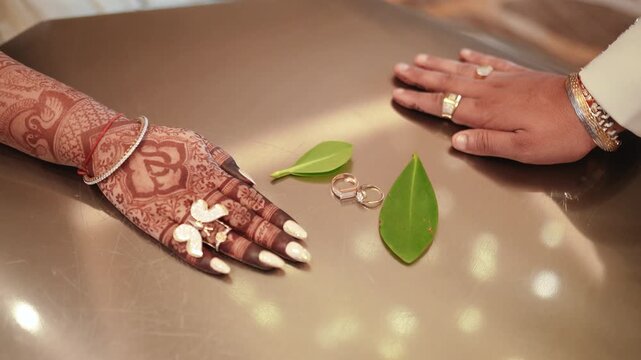 Flat lay of gold wedding rings on leaves with bride's henna hand and groom's hand on table