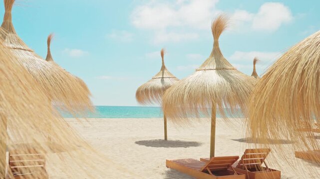 Empty wooden sunbeds and thatched straw umbrellas on a beautiful mediterranean sandy beach with a turquoise ocean