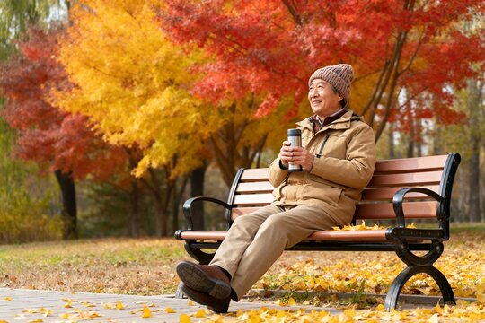 Elderly man sitting on a park bench in autumn, holding a thermos, surrounded by colorful fall foliage.