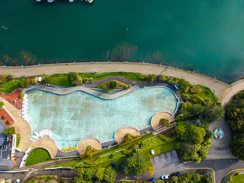 Aerial view of the outdoor swimming pool and surrounding parkland at Bangor Harbour, featuring turquoise water, paved walkways, and lush green gardens in Bangor, Northern Ireland, United Kingdom.
