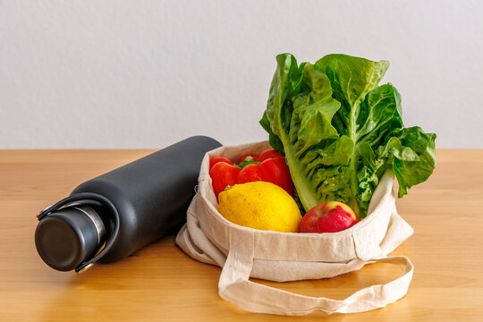Reusable cotton shopping bag with fresh vegetables, lemon and apple next to a black stainless steel water bottle on wooden table, zero waste and sustainable lifestyle concept