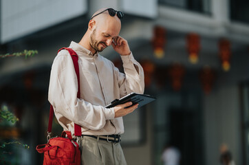 A bald man with a red shoulder bag reads a tablet while standing on an urban street. He looks thoughtful and focused as he checks the mobile device outdoors. © qunica.com
