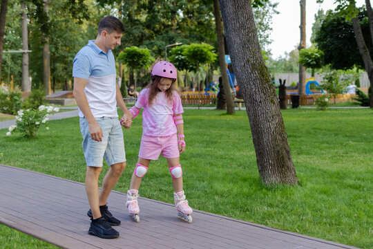 Father holding hands with his little girl in full safety gear learning to rollerblade in a park. Rollerblading and active kid leisure concept
