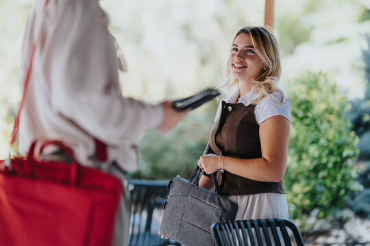 A smiling female customer holds a gray bag while interacting with a staff member holding a payment device. The scene shows a friendly outdoor transaction between customer and service staff.
