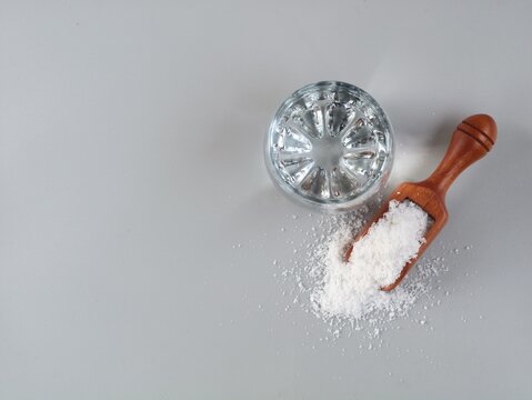 Glass of clear saltwater solution next to a wooden scoop filled with white sea salt on a minimalist grey background. Ideal for health, science, cooking, and natural remedy concepts