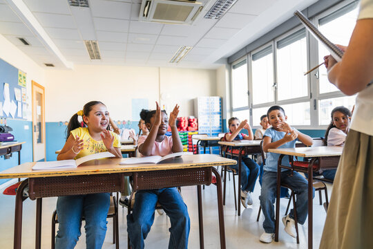 Happy elementary students sitting at desks, clapping hands during an interactive lesson with their teacher. Education concept