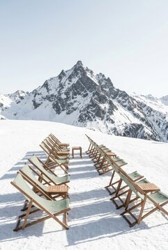 Rows of wooden deck chairs with sage green canvas on snow facing a sharp snow capped alpine peak on a sunny day - Ski resort terrace, winter holiday and apres ski relaxation