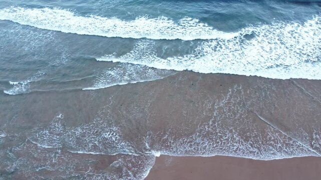 Aerial view of blue ocean waves washing over a dark sandy beach, creating layered foam, wet shoreline textures, and elegant coastal patterns.