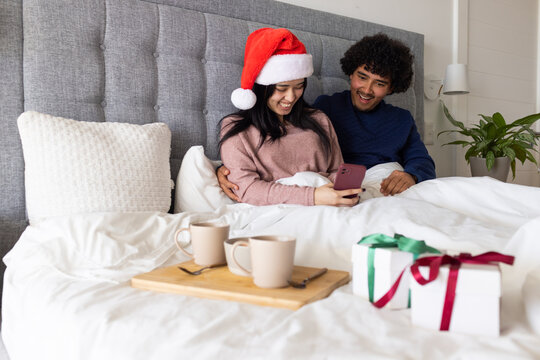 African American couple sitting in bed wearing Santa hat holding smartphone sharing mugs christmas