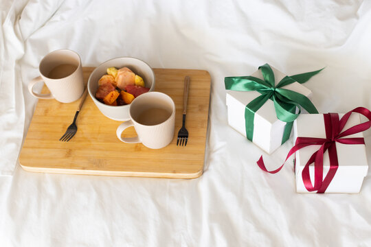 Bamboo tray is holding two ribbed mugs and fruit bowl on white bed, christmas gifts