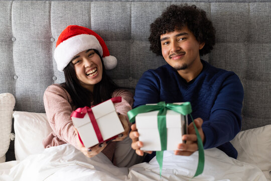 Couple sitting up in bed presenting wrapped christmas gifts wearing Santa hat and navy sweater