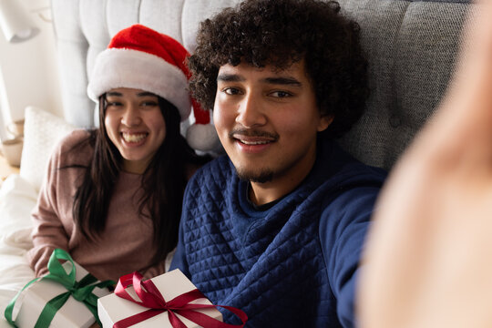 Couple on bed by tufted headboard woman wearing Santa hat man taking christmas selfie holding gifts