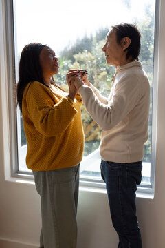 Senior Asian man and African American woman holding eyeglasses at large window in knit sweaters