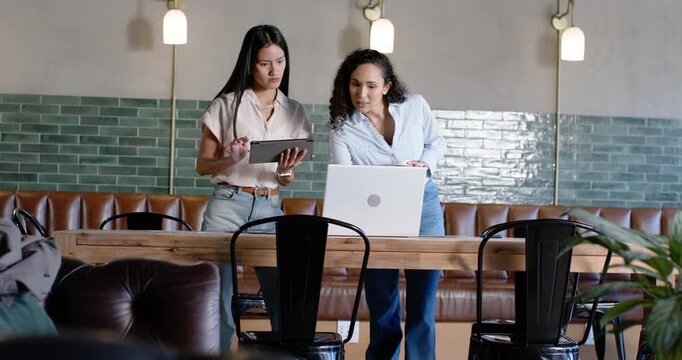 Diverse female coworkers responding to tablet content, scrolling, pointing at laptop in cafe lounge