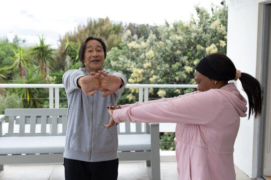 Senior Asian man and woman stretching arms forward on covered balcony near bench, wearing hoodies