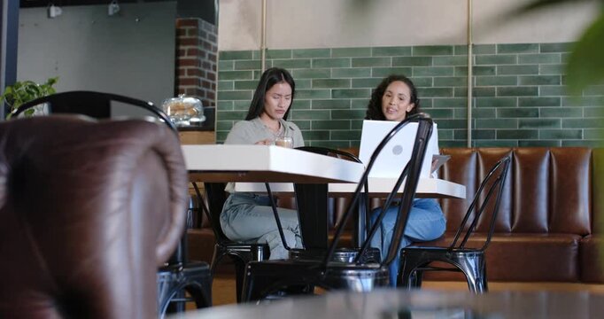 Diverse female friends in light green and white at cafe, right explaining laptop, left leaning in
