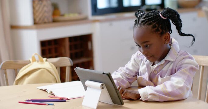 African female child after reaching for pencils tapping tablet on stand in kitchen doing homework