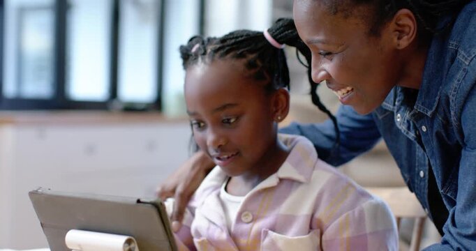 African woman placing guiding hand, leaning in and encouraging youth girl tapping tablet at table
