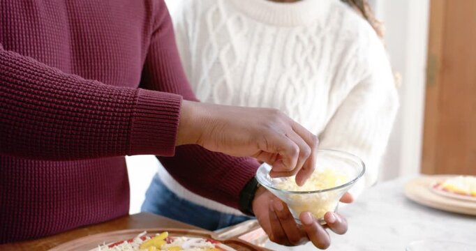 African couple preparing pizza at counter, man sprinkling cheese from bowl while woman tasting