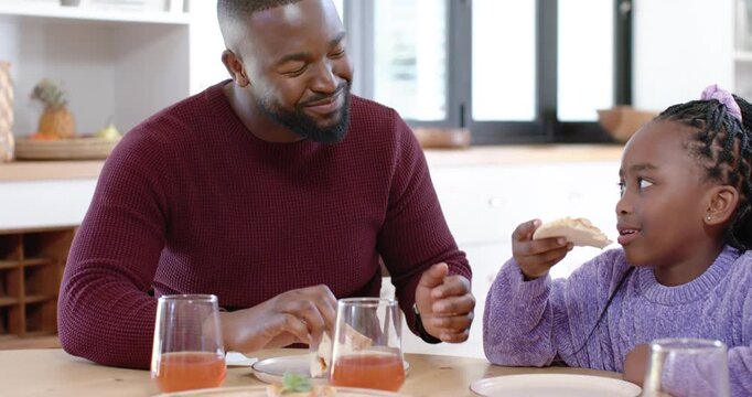 African American father and daughter eating pizza at kitchen table, father leaning, wiping fingers