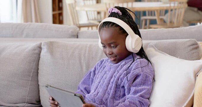 African school-age girl tapping tablet to start app, wearing white headphones, smiling on gray sofa