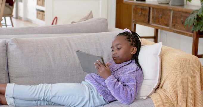 African youth female reclining on gray sofa in purple sweater glancing refocusing studying tablet