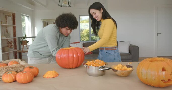 Asian woman and African American man carving and scooping pumpkin into bowls at home for halloween