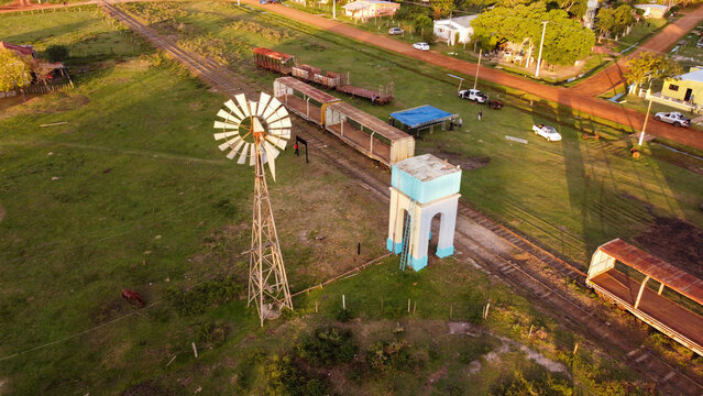 Aerial view of a rural landscape featuring a vintage windmill, a blue and white water tower, and old train cars resting on tracks in Guaviravi, Corrientes Province, Argentina.