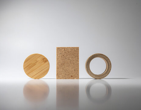 studio still life of round wooden disk, rectangular cork block, and recycled cardboard ring arranged on reflective white surface with soft gradient lighting