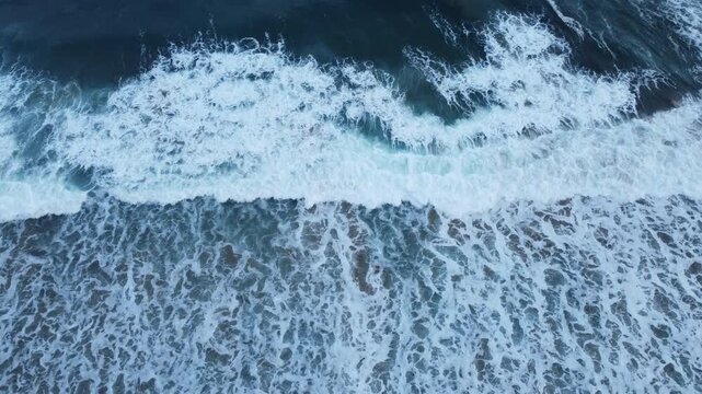 Top-down drone footage of white ocean foam spreading across blue-gray water, forming delicate branching patterns and fluid natural textures.