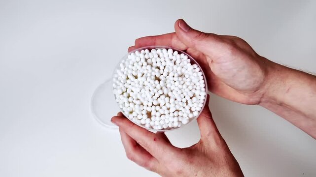 A pair of hands carefully holds a clear plastic container filled with numerous small white spherical objects against a plain white background, presenting the items for view