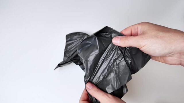 A person's hands are shown holding and manipulating a crumpled piece of black plastic material against a plain white surface, demonstrating the action of handling trash or refuse