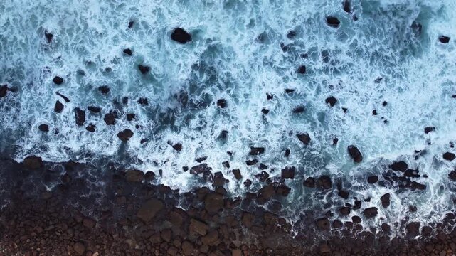 Overhead view of foamy waves washing across dark coastal rocks, creating a high-contrast pattern of white surf, blue water, and rugged shoreline.