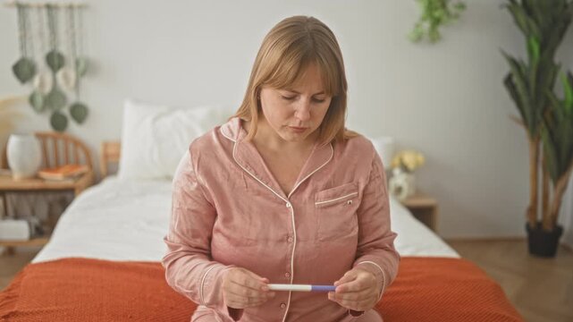 Woman in pink pajamas holding a pregnancy test, hands and gaze fixed on the result while seated on a bed in a bedroom; anxiety.