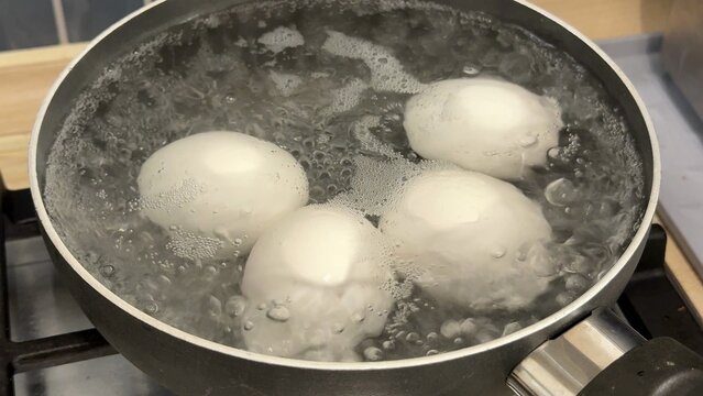 Several whole white eggs gently float in a large pot of boiling water on a stove top, the water surface showing many bubbles and steam creating a cooking scene