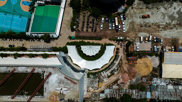 Aerial view of a building complex with a semi-circular white canopy, adjacent sports courts, and parking areas in the urban landscape of Dhaka, Dhaka Division, Bangladesh.