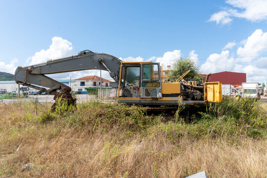 Old tracked hydraulic excavator abandoned in tall grass and vegetation near an industrial area. Extended boom, hydraulic cylinders, exposed engine components and weathered metal illustrate heavy const