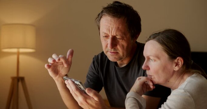 Mature couple at home reviewing something on a mobile phone together, worried expressions, concept of shared concern and communication, soft indoor lighting. 4K.