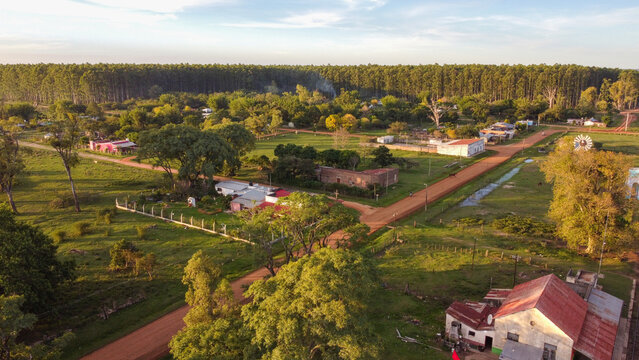 Aerial view of the rural town with dirt roads, scattered houses, lush green trees, and a dense forest backdrop during golden hour in Guaviravi, Corrientes Province, Argentina.